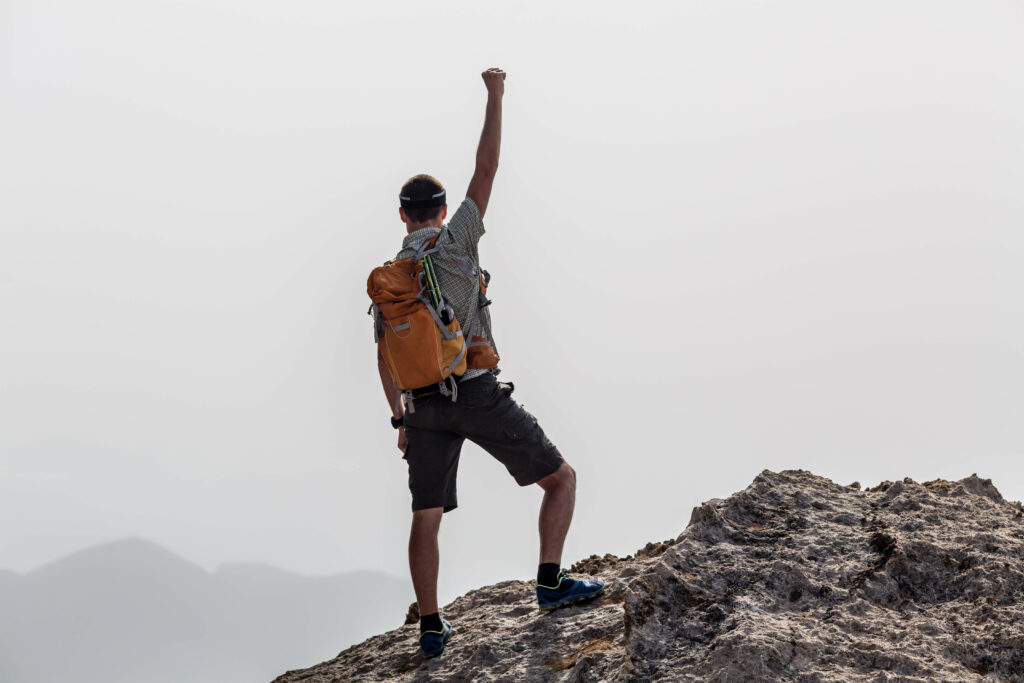 man at the top of a mountain reaching his arm up in celebration